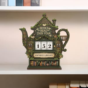 May include: A whimsical, green teapot-shaped bookshelf with the text "BOOKS READ THIS YEAR" and "SOPHIA'S LIBRARY." It features a counter displaying "152" and is adorned with miniature books, windows, and foliage. The shelf is made of wood and sits on a white shelf.
