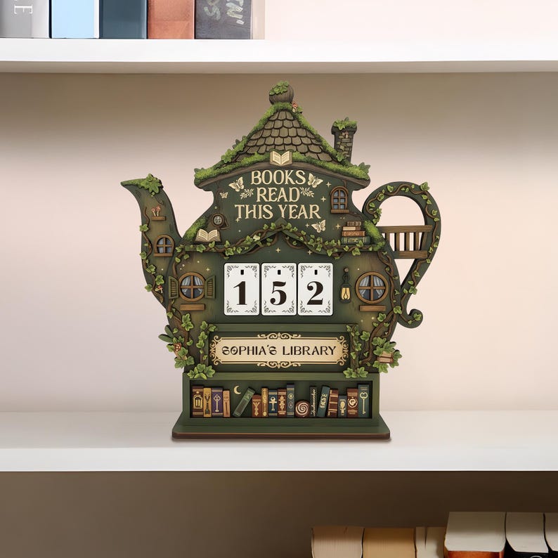 May include: A whimsical, green teapot-shaped bookshelf with the text "BOOKS READ THIS YEAR" and "SOPHIA'S LIBRARY." It features a counter displaying "152" and is adorned with miniature books, windows, and foliage. The shelf is made of wood and sits on a white shelf.