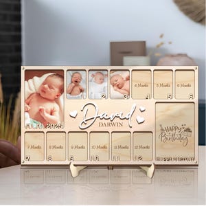 Puede incluir: Un marco de fotos de madera con un calendario para el primer año de un bebé. El marco tiene 12 ranuras para fotos y un espacio para el nombre y la fecha de nacimiento del bebé. El marco está decorado con corazones y las palabras "Feliz cumpleaños".