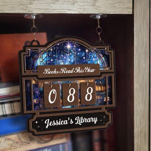 Könnte beinhalten: Ein hölzerner Bücherzähler mit dem Text "Books Read This Year" und "Jessica's Library". Der Zähler zeigt "088" mit einem Sternennacht-Design im Hintergrund an. Der Zähler hängt an einem Regal.