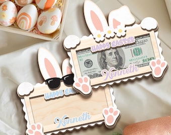 May include: Two wooden bunny-shaped picture frames with a scalloped edge. The frames are light brown with pink bunny ears and paw prints. The top frame has a dollar bill inside with the text "Happy Easter Kenneth". The bottom frame has the text "Happy Easter Kenneth".