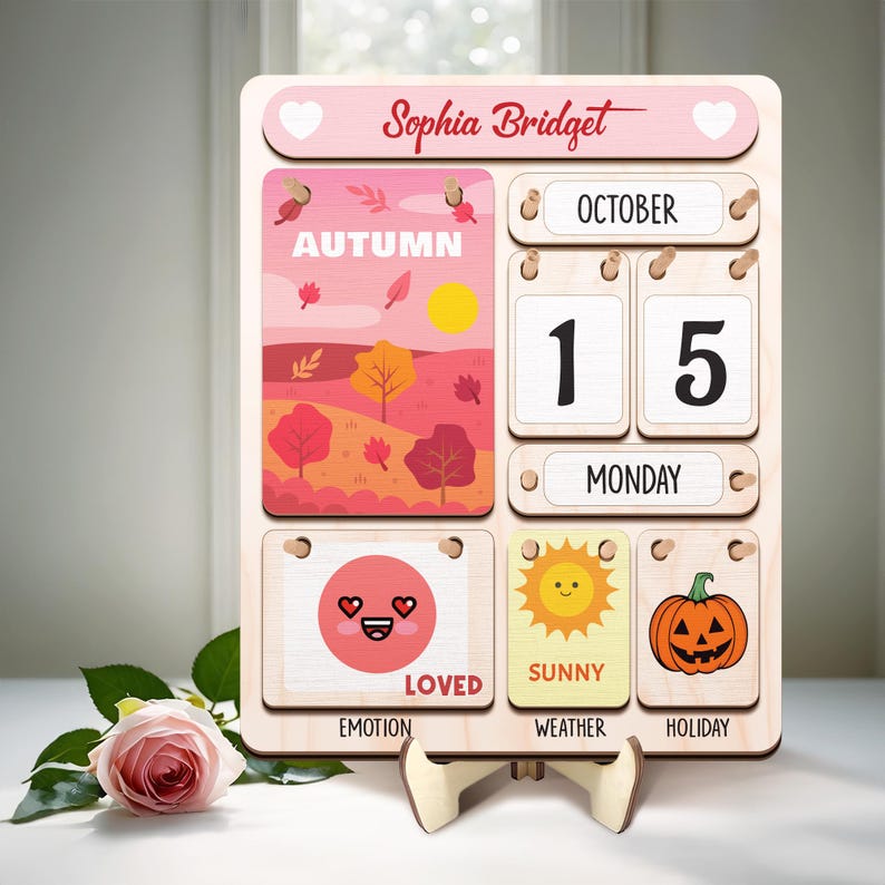 May include: A pink wooden calendar with the name "Sophia Bridget" at the top. The calendar has a date display, a day of the week display, and a section for the month, weather, emotion, and holiday. The current date is October 15th, and the day of the week is Monday. The month is Autumn, the weather is sunny, the emotion is loved, and the holiday is a pumpkin.