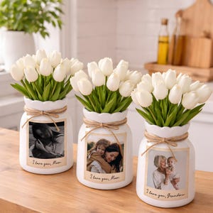 Puede incluir: Tres jarrones de cerámica blanca con tulipanes blancos y fotos familiares. Cada jarrón tiene un lazo de cuerda y el texto "I love you, Mom" o "I love you, Grandma". Los jarrones están sobre una superficie de madera.