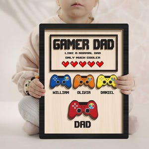 May include: A wooden sign with the text "Gamer Dad" and a pixelated heart design. The sign features four colourful game controllers with names below each, and a larger controller at the bottom with the word "Dad".