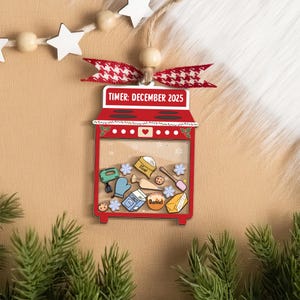 May include: A red and white Christmas ornament shaped like an oven, with the text "TIMER: DECEMBER 2025". The ornament features baking-themed illustrations inside, including a mixer, flour, and cookies. It is decorated with a red and white bow.