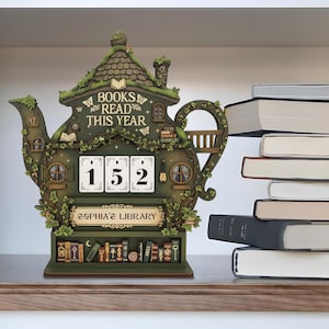 May include: A whimsical, green teapot-shaped bookshelf calendar with the text "BOOKS READ THIS YEAR" and the number "152". The calendar has a miniature library at the base and is decorated with vines and butterflies.