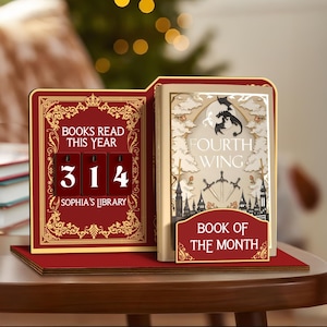 Puede incluir: Un expositor de libros de madera roja y dorada con el texto "BOOKS READ THIS YEAR" y el número "314". Un libro titulado "FOURTH WING" se muestra junto a él, con un letrero que dice "BOOK OF THE MONTH".