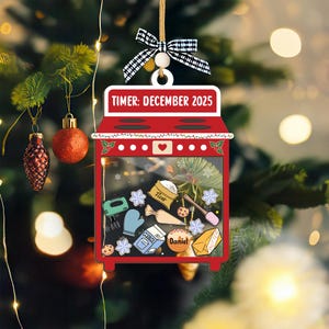 May include: A red oven-shaped Christmas ornament with the text "TIMER: DECEMBER 2025". The ornament features baking-themed illustrations inside, including a mixer, snowflakes, and a bag of flour. It is suspended by a string with a black and white checkered bow.