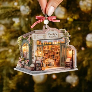 May include: A miniature bookstore ornament with the text "Emily's Bookstore." The ornament features a detailed facade with a striped awning, a guitar, and a piano. The interior is filled with bookshelves and soft lighting, and is suspended by a red and white ribbon.