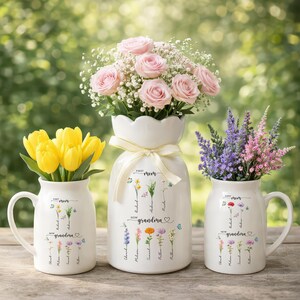 Pode incluir: Três vasos de cerâmica branca com arranjos florais. O vaso central contém rosas cor-de-rosa, o vaso esquerdo tulipas amarelas e o vaso direito flores roxas e cor-de-rosa. Cada vaso tem as palavras "First Mom Now Grandma" e ilustrações florais.
