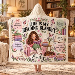 May include: A cream-colored hooded blanket featuring the text 'THIS IS MY READING BLANKET' and 'Emily'. The design includes a cartoon girl reading in a chair, a dachshund, stacks of books, floral accents, and phrases like 'Just one more chapter'.