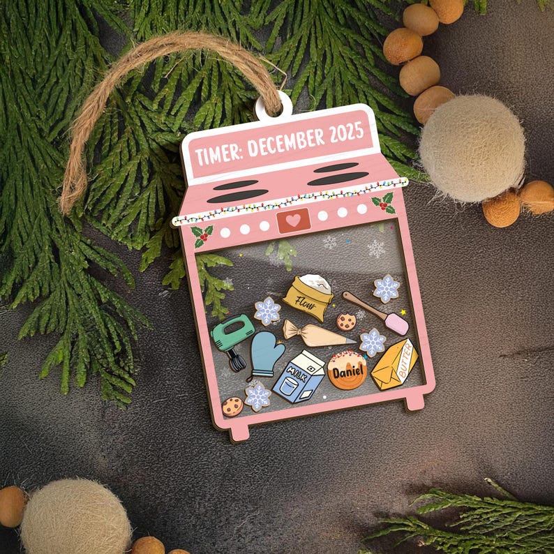 May include: A pink wooden ornament shaped like an oven, with the text "TIMER: DECEMBER 2025". Inside the clear window are baking-themed illustrations, including flour, milk, and cookies. The ornament is strung with twine.