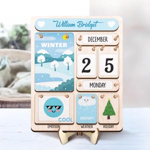 May include: A wooden calendar with a winter theme. The calendar features a blue and white winter scene, a snowman, a Christmas tree, and a cloud with snowflakes. The date is December 25th, and the day is Monday. The calendar also includes the words "Winter", "Cool", "Snowy", and "Holiday".