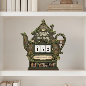 May include: A whimsical, green teapot-shaped bookshelf decor piece with the words "BOOKS READ THIS YEAR." It features a counter displaying "152" and the text "SOPHIA'S LIBRARY." The design includes miniature books and a charming, fairy-tale aesthetic.