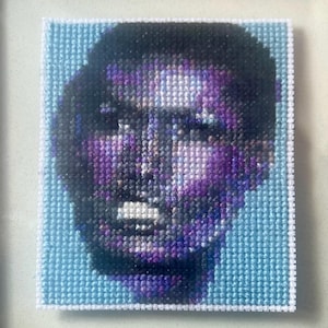 May include: A framed cross-stitch portrait of a person with a purple and blue face against a light blue background.