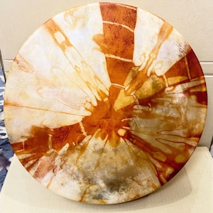 May include: A circular drum with a stretched skin head featuring a tie-dye pattern in shades of burnt orange, deep rust, and cream. The design radiates from the center toward the edges in a sunburst effect with organic, fluid lines.