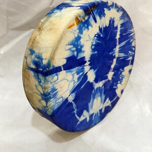 May include: A circular frame drum featuring a tie-dye pattern in shades of deep blue and cream. The drum head displays a radial, sunburst-like design with organic, flowing lines created by the dye, while the side frame shows a natural tan texture.