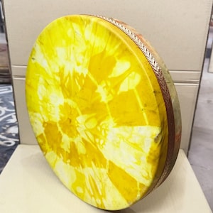 May include: A round frame drum featuring a vibrant yellow and white tie-dye pattern on the drumhead. The circular wooden frame is wrapped in a decorative brown trim with a repeating leaf-like pattern along the outer edge of the instrument.