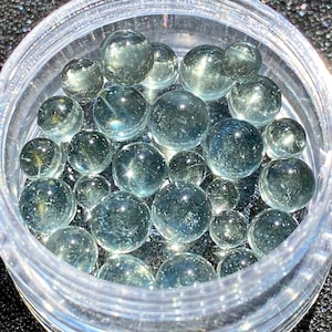 May include: A clear plastic container filled with many small, round, light blue glass marbles.