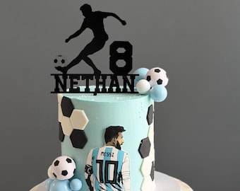 May include: A light blue cake decorated with black and white soccer ball patterns, featuring a cutout of a soccer player wearing a number 10 jersey with the name Messi, and a black cake topper displaying the name Nethan and the number 8.
