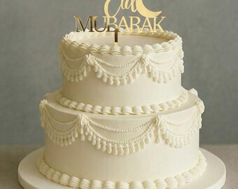 May include: A two-tiered white cake with intricate buttercream frosting details. The cake is decorated with a gold "Eid Mubarak" cake topper. The cake sits on a white cake stand, perfect for a celebration.