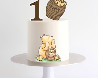 May include: A white cake decorated with a Winnie the Pooh theme. The cake features a brown number 1, a cartoon image of Winnie the Pooh, and a honey pot with the word "Hunny". The cake is on a white cake stand.