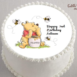 May include: A white frosted cake with a Winnie the Pooh themed edible image. The image features Winnie the Pooh with a honey pot, bees, and the text "Happy 2nd Birthday Autumn". The cake is decorated with white frosting around the edges.