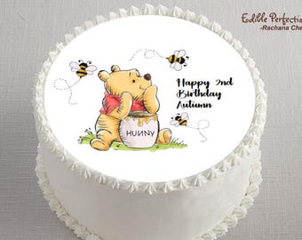 May include: A white frosted cake with a Winnie the Pooh themed edible image. The image features Winnie the Pooh with a honey pot, bees, and the text "Happy 2nd Birthday Autumn". The cake is decorated with white frosting around the edges.