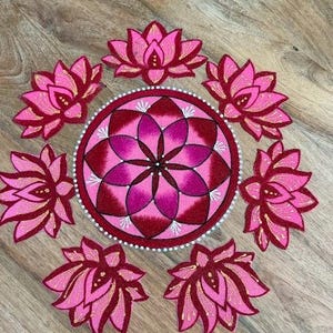 May include: A circular rangoli design featuring a central mandala with layered pink and deep red petals, surrounded by six individual lotus flower motifs in matching shades of pink, red, and gold, all arranged in a symmetrical floral pattern.