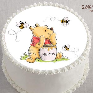May include: A white cake decorated with a Winnie the Pooh image. The image features a yellow bear with a red shirt, sitting next to a honey pot with the word "HUUNY". Two bees fly around the bear. The cake has white frosting.