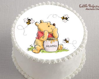 May include: A white cake decorated with a Winnie the Pooh image. The image features a yellow bear with a red shirt, sitting next to a honey pot with the word "HUUNY". Two bees fly around the bear. The cake has white frosting.