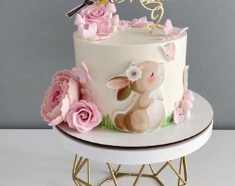 May include: A white cake decorated with a pink bunny, pink flowers, a yellow bird, and a gold name topper that says "Milana". The cake is on a white cake stand with a gold geometric base.
