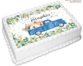 May include: A rectangular cake with white frosting and a decorative border. The cake is topped with an edible image featuring a blue truck carrying pumpkins, surrounded by greenery. Text reads "a sweet little pumpkin is on the way."
