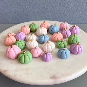 May include: A collection of miniature pumpkins in various pastel colors, including pink, green, blue, white, and purple. Each pumpkin has a gold-colored stem. The pumpkins are arranged on a white marble-like surface.