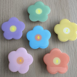 May include: Six flower-shaped objects in pastel colours: green, yellow, blue, pink, purple, and coral. Each flower has a central circle with a number from 1 to 6. The objects are arranged on a light wooden surface.