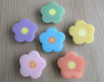 Può includere: Sei oggetti a forma di fiore dai colori pastello: verde, giallo, blu, rosa, viola e corallo. Ogni fiore ha un cerchio centrale con un numero da 1 a 6. Gli oggetti sono disposti su una superficie di legno chiaro.
