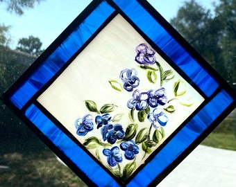 May include: A stained glass window featuring a diamond-shaped frame with a blue border. The center of the window is a white background with a painted design of purple flowers and green leaves.
