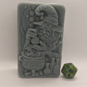 May include: A gray, rectangular soap bar with a detailed carving of a wizard holding a cauldron and a potion bottle. The wizard is wearing a pointed hat and has a long beard. The soap bar is surrounded by bubbles.