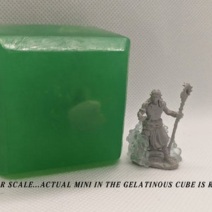 May include: A green gelatinous cube with a miniature figure inside. The miniature is a grey figure with a staff and is standing on a base. The cube is about 2 inches tall. "MINI FOR SCALE...ACTUAL MINI IN THE GELATINOUS CUBE IS RANDOM" is written below the image.