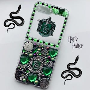 Könnte beinhalten: Eine transparente Handyhülle, verziert mit grünen und schwarzen Verzierungen, mit einem Slytherin-Wappen und den Worten "Harry Potter". Die Hülle ist mit grünen Edelsteinen, Perlen und einem kleinen Schlangendesign verziert.