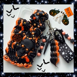 Peut inclure: Une coque de t&eacute;l&eacute;phone sur le th&egrave;me d'Halloween noire et orange avec un cr&acirc;ne, des chauves-souris et une toile d'araign&eacute;e. La coque a un charme d&eacute;tachable avec un tourbillon noir et orange, des &eacute;toiles noires et des perles roses.