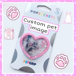 May include: A pink heart-shaped phone grip with a clear window showing a cat inside. The grip is decorated with glitter and stars. The packaging says "POPSOCKETS Custom Pet Image" and "POPGRIP".