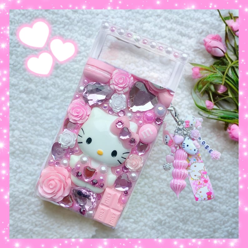May include: A pink and white Hello Kitty phone case with rhinestones, pink roses, and a pink and white chocolate bar. The case has a clear window on the front and a pink and white Hello Kitty charm attached to it.