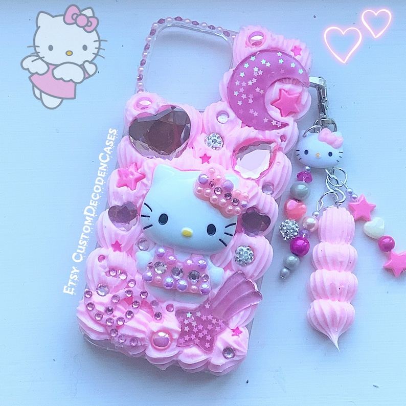 May include: A pink Hello Kitty phone case with a 3D design featuring Hello Kitty, stars, hearts, and a crescent moon. The case has a pink and white beaded charm attached.