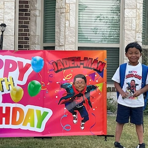 May include: A colorful banner with the text "Happy 6th Birthday" and a cartoon superhero character with the text "Jaden-Man". A young boy wearing a white t-shirt with the text "Happy Jaden-Man Birthday" is standing in front of the banner.