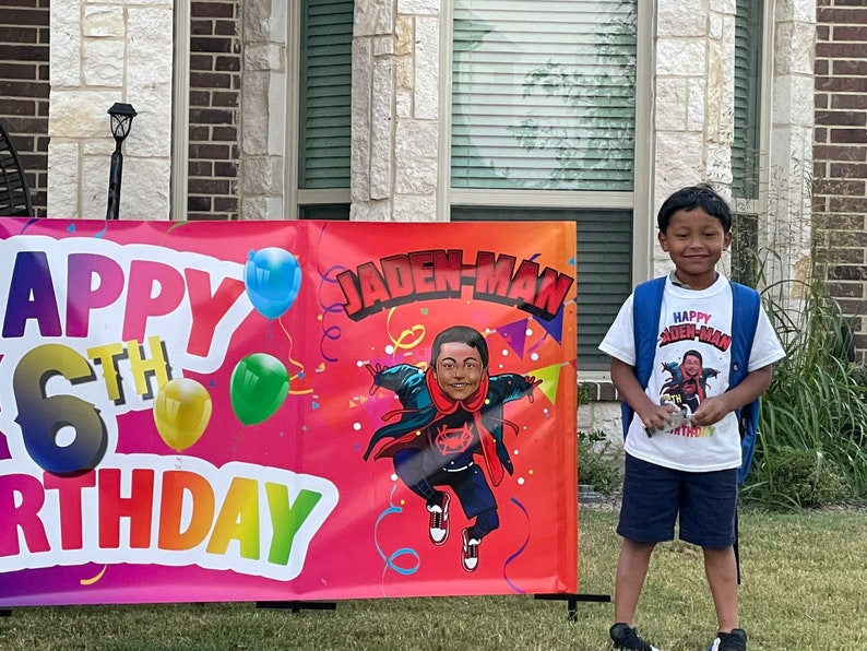 May include: A colorful banner with the text "Happy 6th Birthday" and a cartoon superhero character with the text "Jaden-Man". A young boy wearing a white t-shirt with the text "Happy Jaden-Man Birthday" is standing in front of the banner.