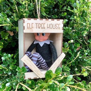 May include: A wooden elf tree house ornament with the words "ELF TREE HOUSE" etched on the top. Inside, an elf doll is seated, wearing a black outfit and striped socks. The ornament hangs from a natural twine string.