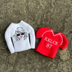 May include: Two miniature felt shirts. One is white with a black and red graphic of a woman wearing sunglasses. The other is red with white trim and the name "KELCE" and the number "87" printed on it.