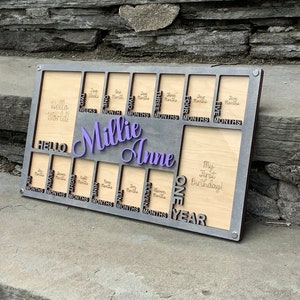 May include: A wooden photo frame with a gray border and a cutout design for photos. The frame is labeled with the months of the year and includes the text "Hello World!" and "My First Birthday!" The frame is personalized with the name "Millie Anne".