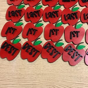 May include: A collection of red wooden apple ornaments with black text. The text on the apples includes "100th", "Last", "1st", and "Best".